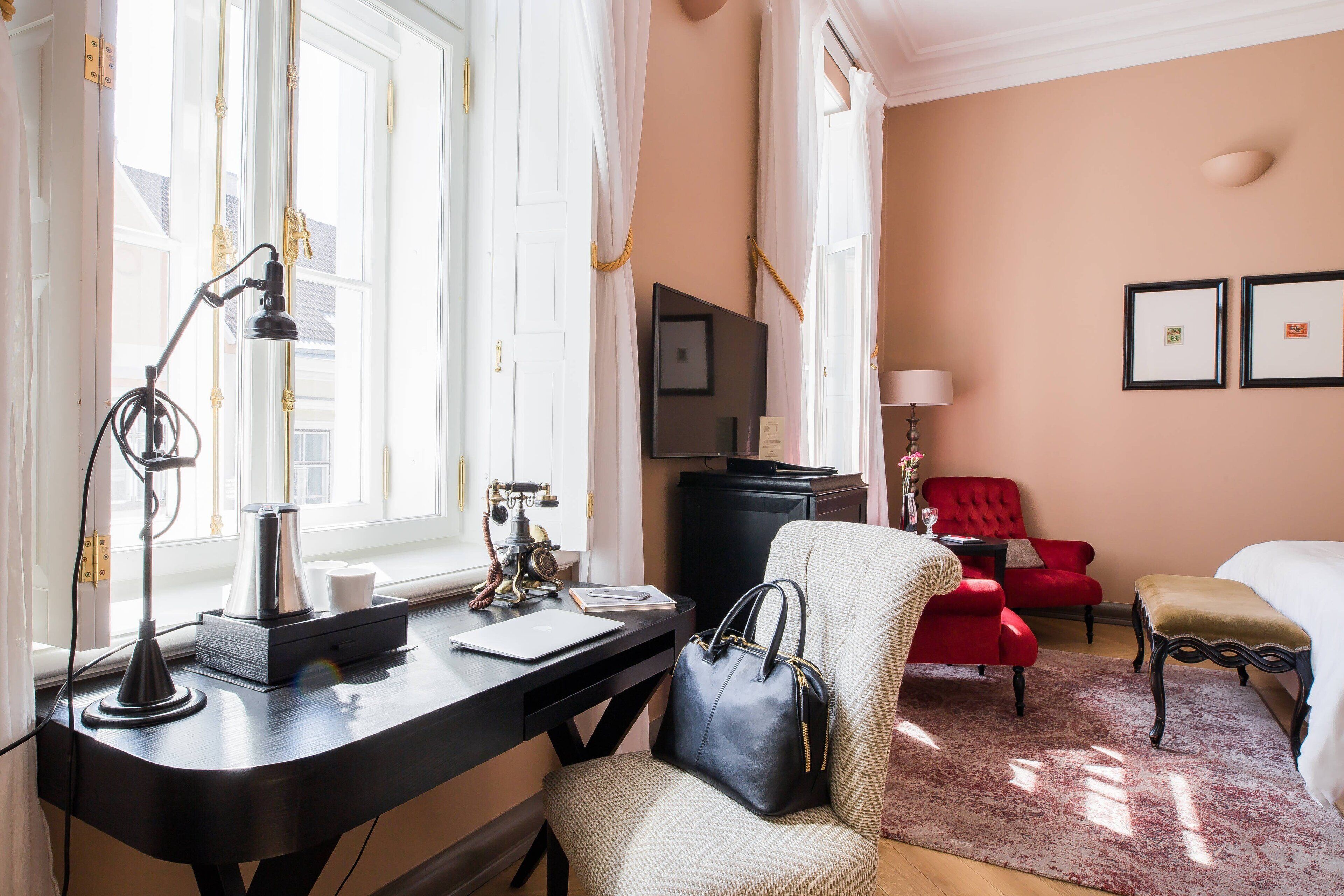 Habitación de hotel con escritorio, un sillón rojo, un sillón blanco, cama y televisión.