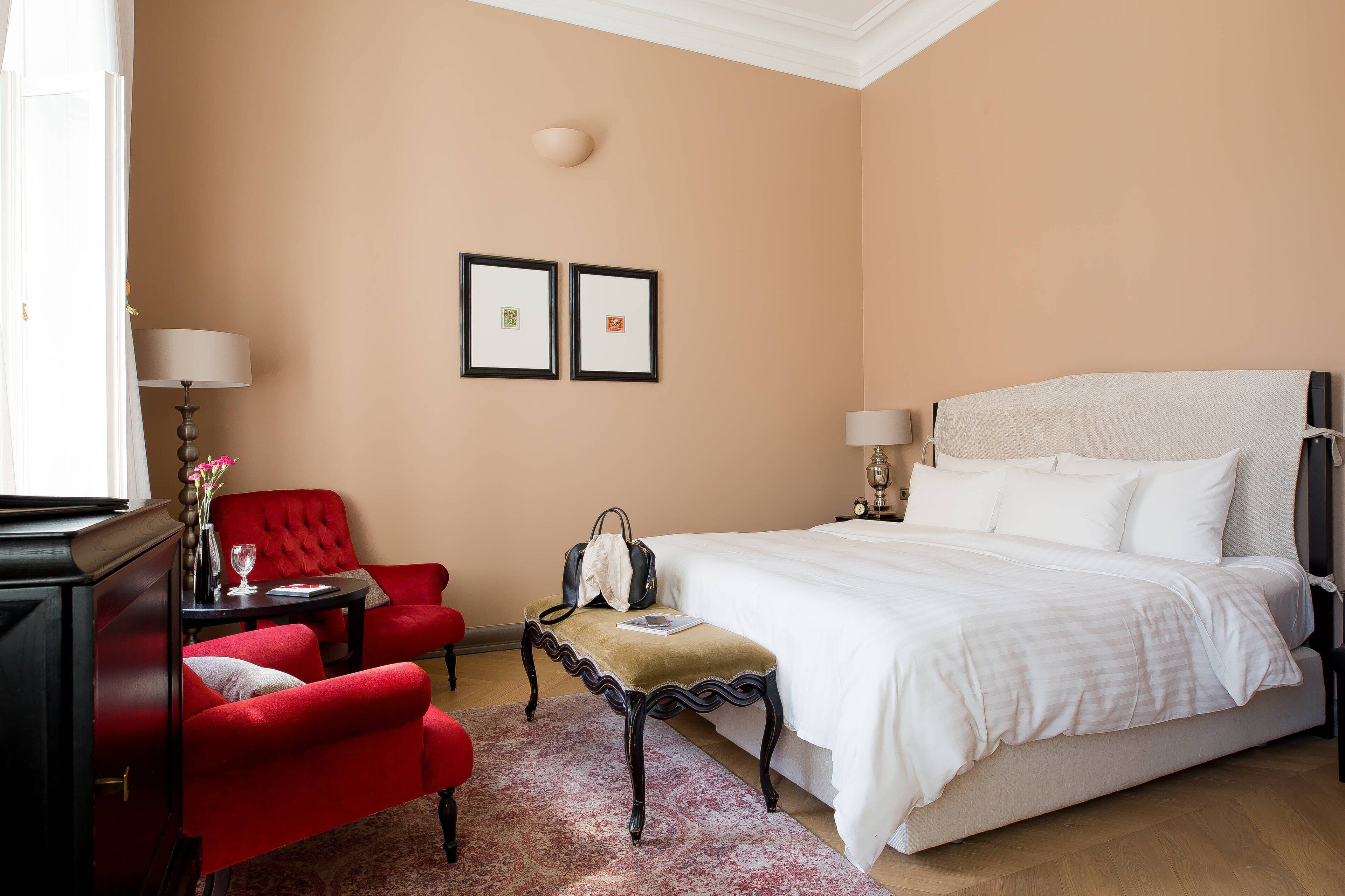 Een hotelkamer met een groot bed, een rode fauteuil, een klein tafeltje met een lamp en twee ingelijste foto's aan de muur.