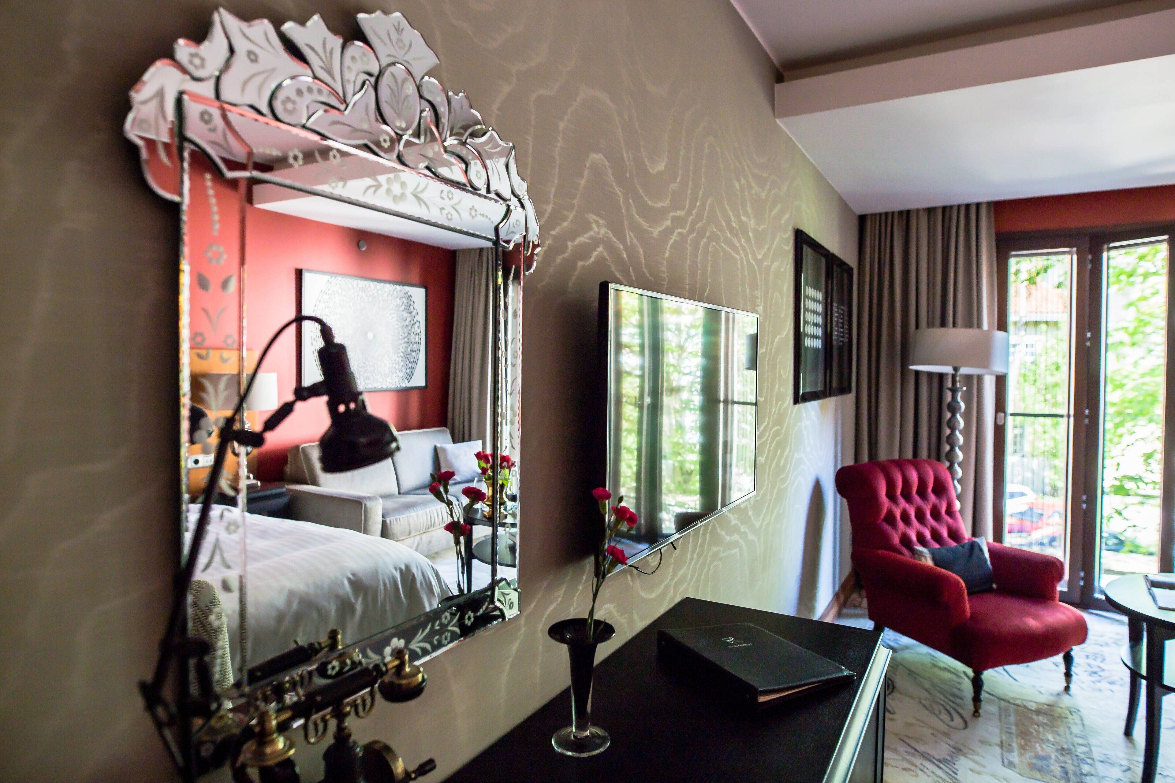 Habitación de hotel con cama, un sillón rojo, escritorio, espejo y ventana con vistas a zonas verdes.