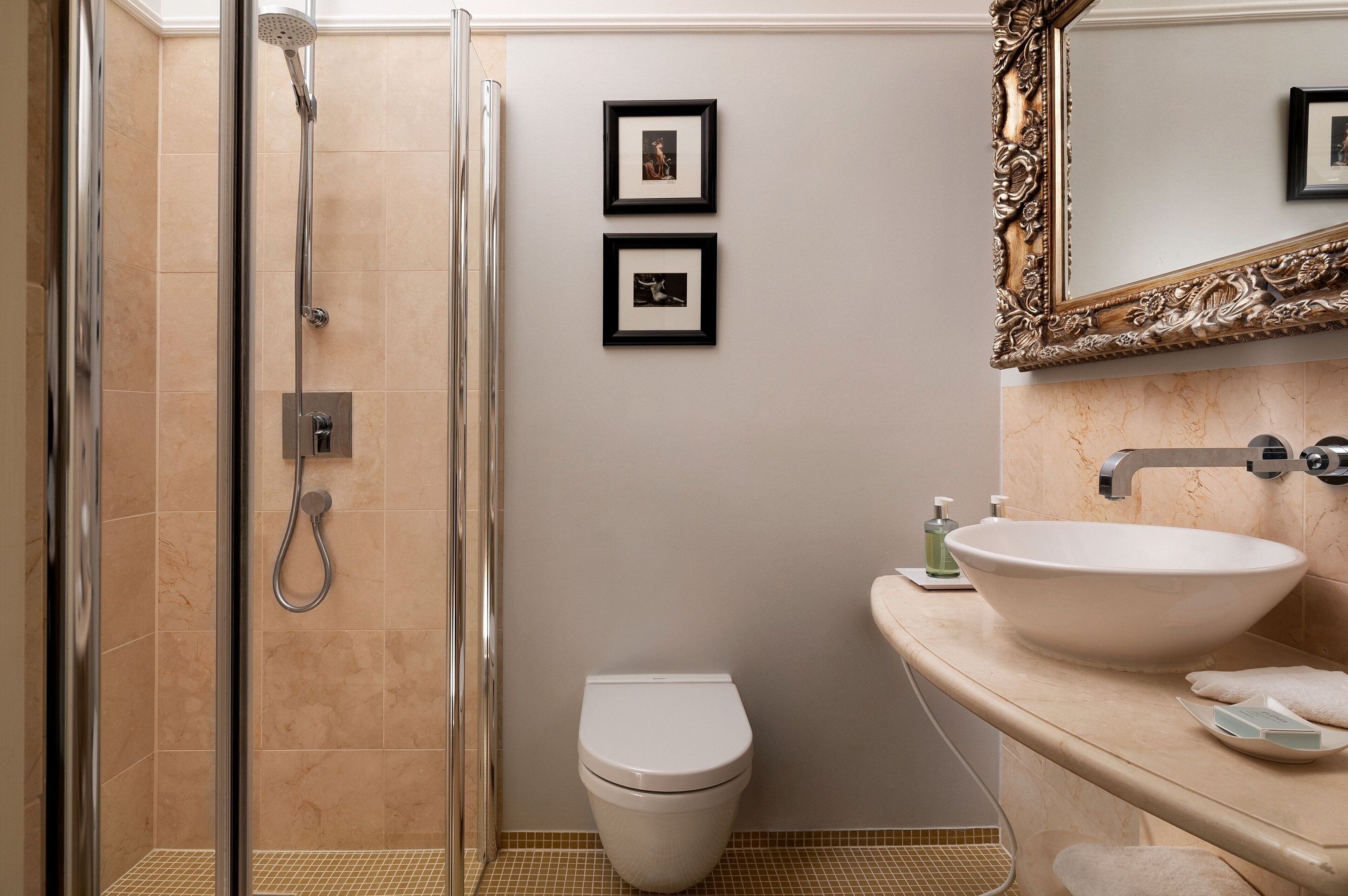 A bathroom with a glass shower enclosure, a toilet, a sink with a marble countertop, and two framed pictures on the wall.