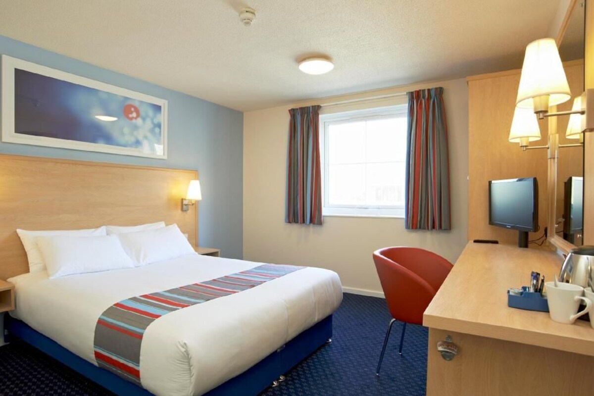 A hotel room with a bed, desk, chair, television, and a painting on the wall.