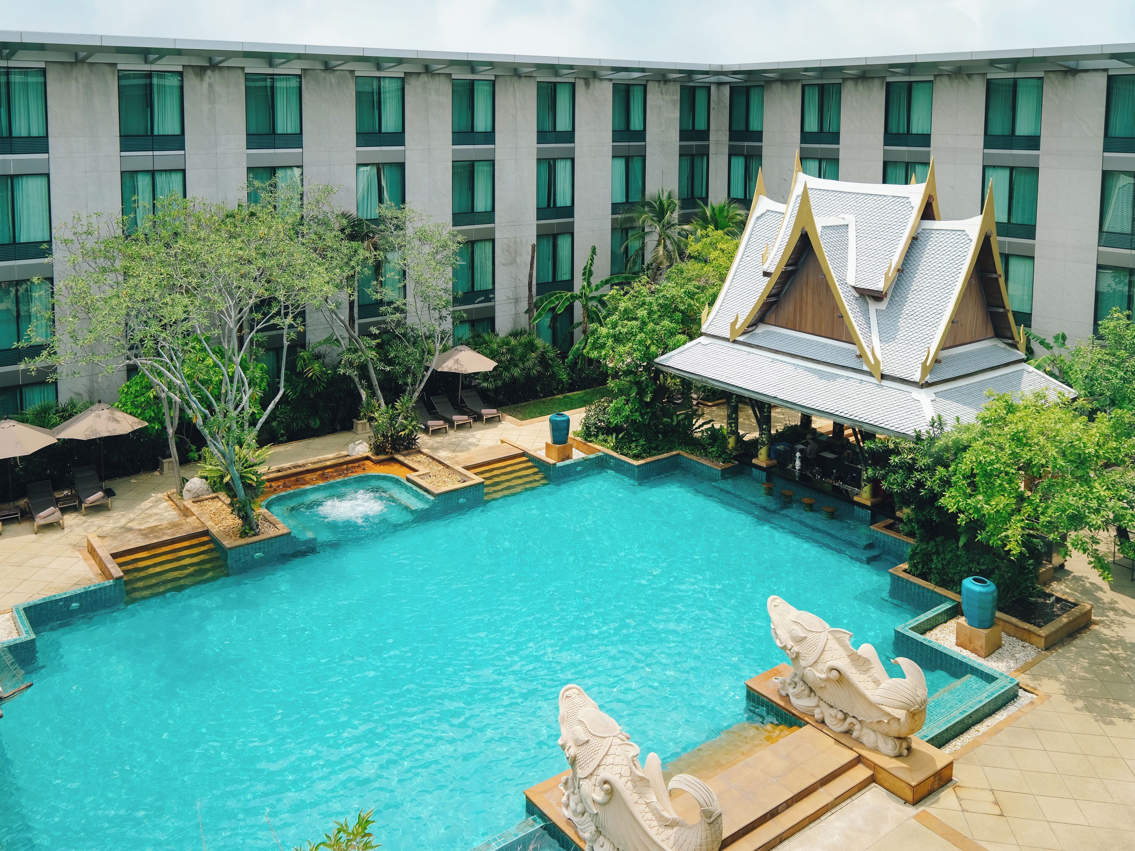A swimming pool with a traditional structure, surrounded by greenery and a modern building with large windows.