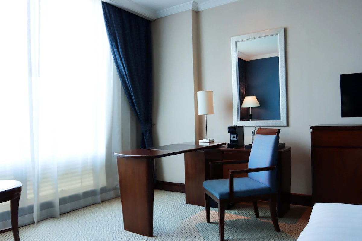 A hotel room with a desk, chair, lamp, and a large mirror on the wall.