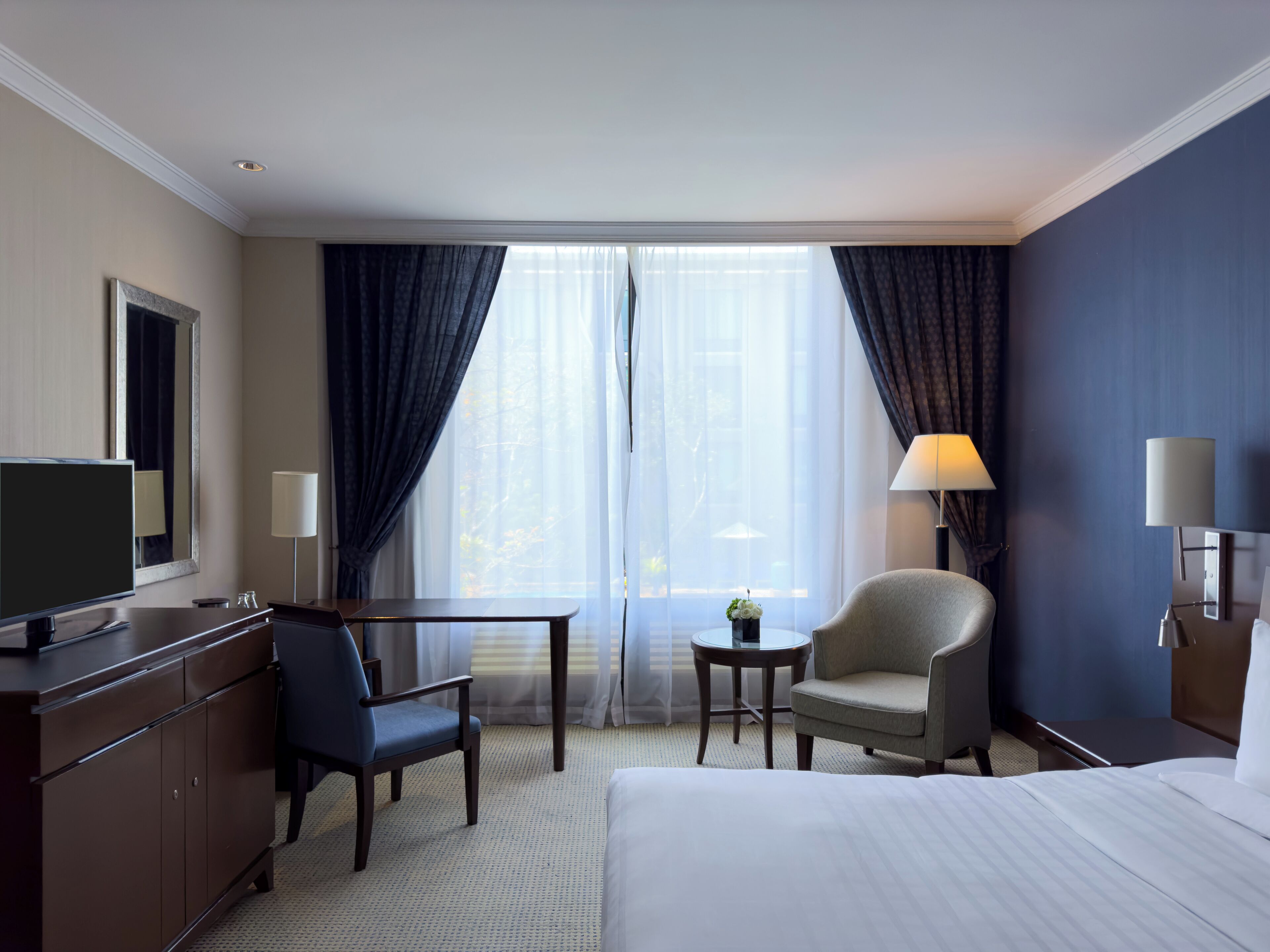 Ein Hotelzimmer mit Bett, Schreibtisch, Stuhl, Fernseher und einem Fenster mit Vorhängen.