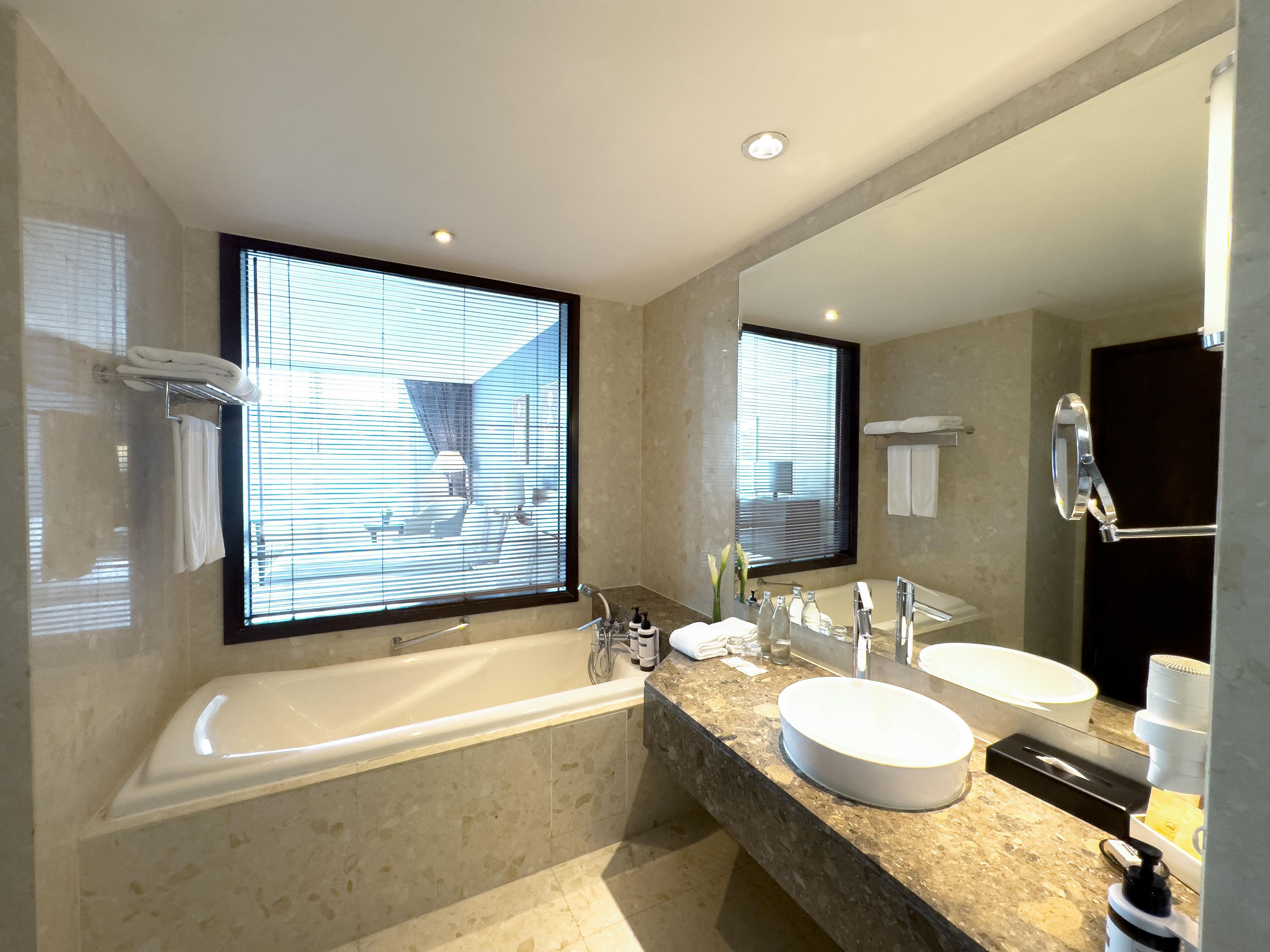 A modern bathroom with a bathtub, two sinks, a large mirror, and a window with blinds.