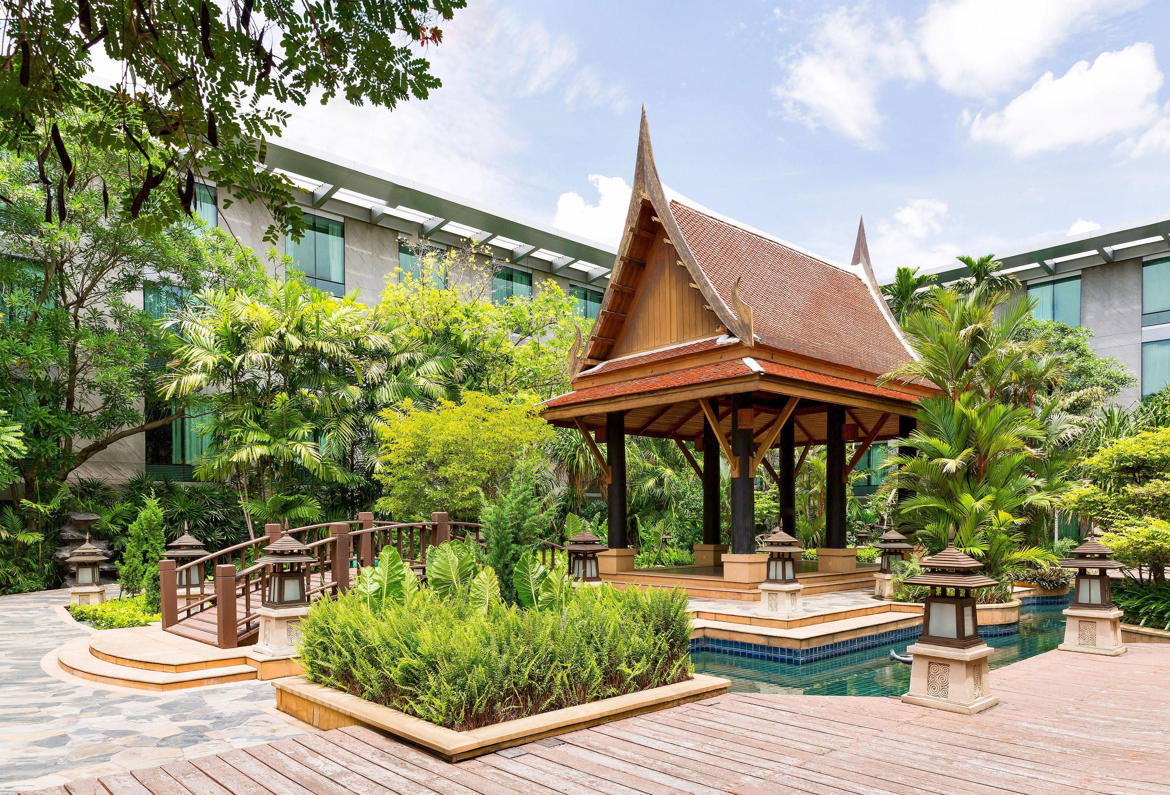 A traditional wooden pavilion with a tiled pool, surrounded by lush greenery and a modern building in the background.