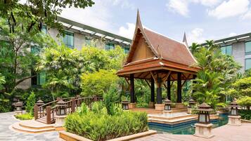 A traditional wooden pavilion with a tiled pool, surrounded by lush greenery and a modern building in the background.