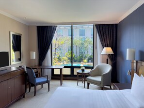 A hotel room with a bed, a desk, a chair, a TV, and a view of a swimming pool and trees.