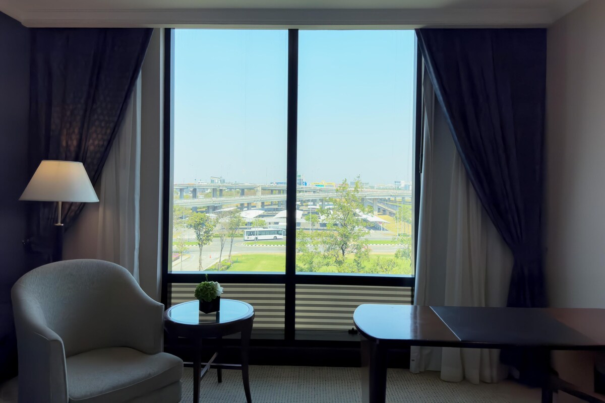 A hotel room with a large window, a chair, a small table with a plant, and a desk.