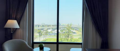 A hotel room with a large window, a chair, a small table with a plant, and a desk.