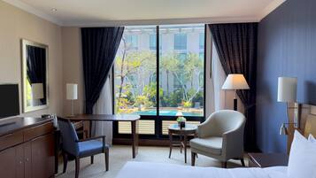 Habitación de hotel con cama, dos sillas, escritorio, televisor y ventana grande con vista a una piscina y vegetación.