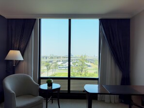 A hotel room with a large window, a chair, a small table with a plant, and a desk.