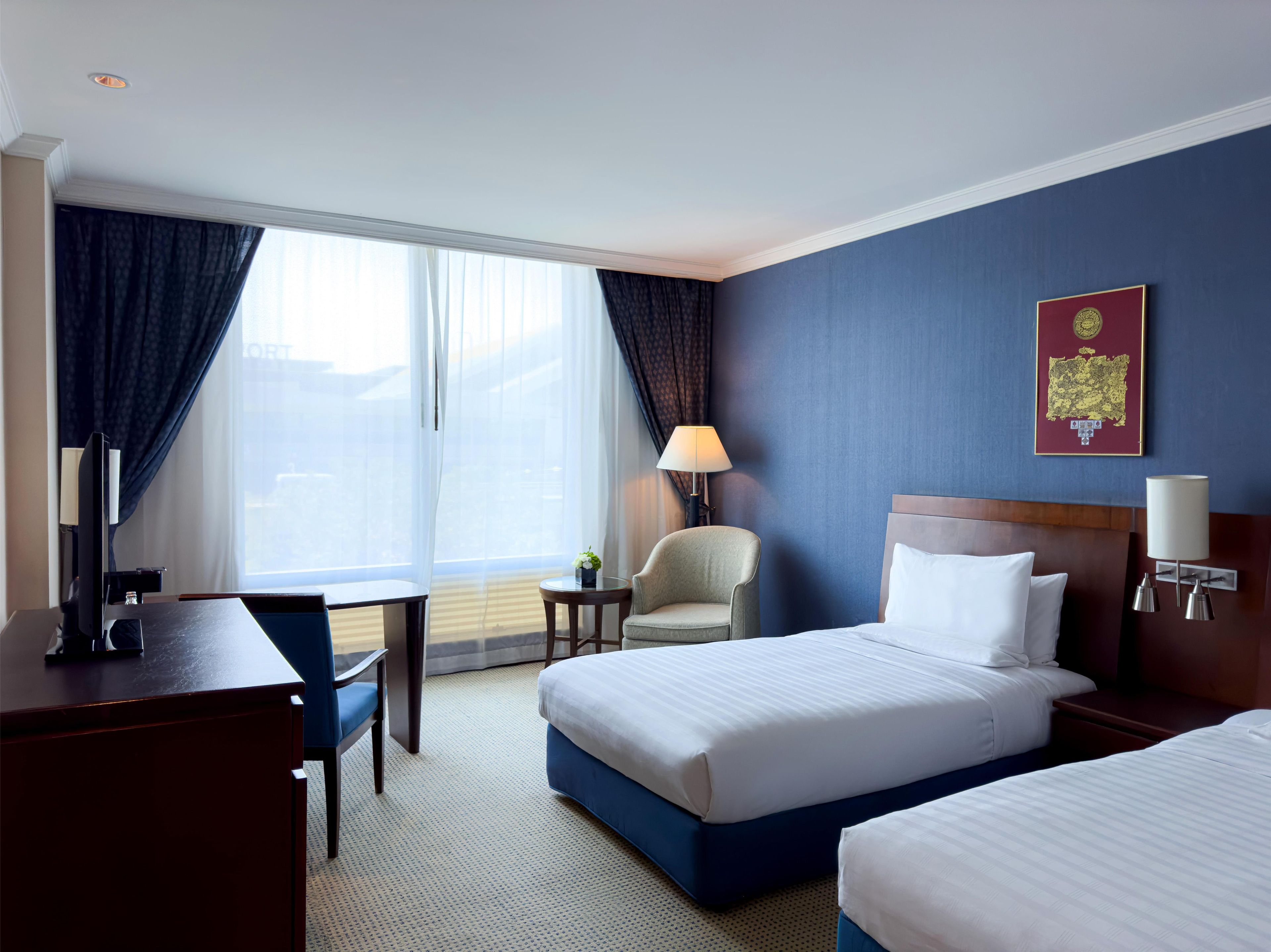 Ein Hotelzimmer mit zwei Betten, einem Schreibtisch, einem Sessel und einem großen Fenster mit Vorhängen.