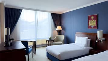 A hotel room with two beds, a desk, a chair, a lamp, and a large window with curtains.