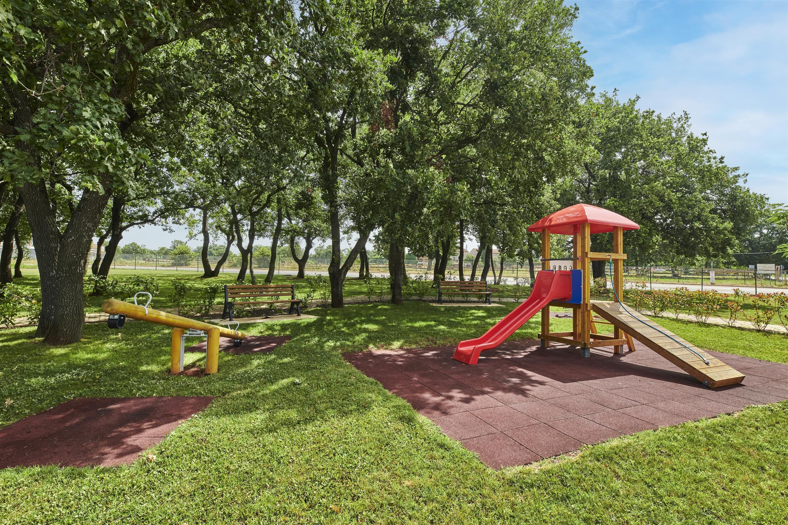 Ein Spielplatz mit Rutsche und Wippe, umgeben von Bäumen und Bänken.