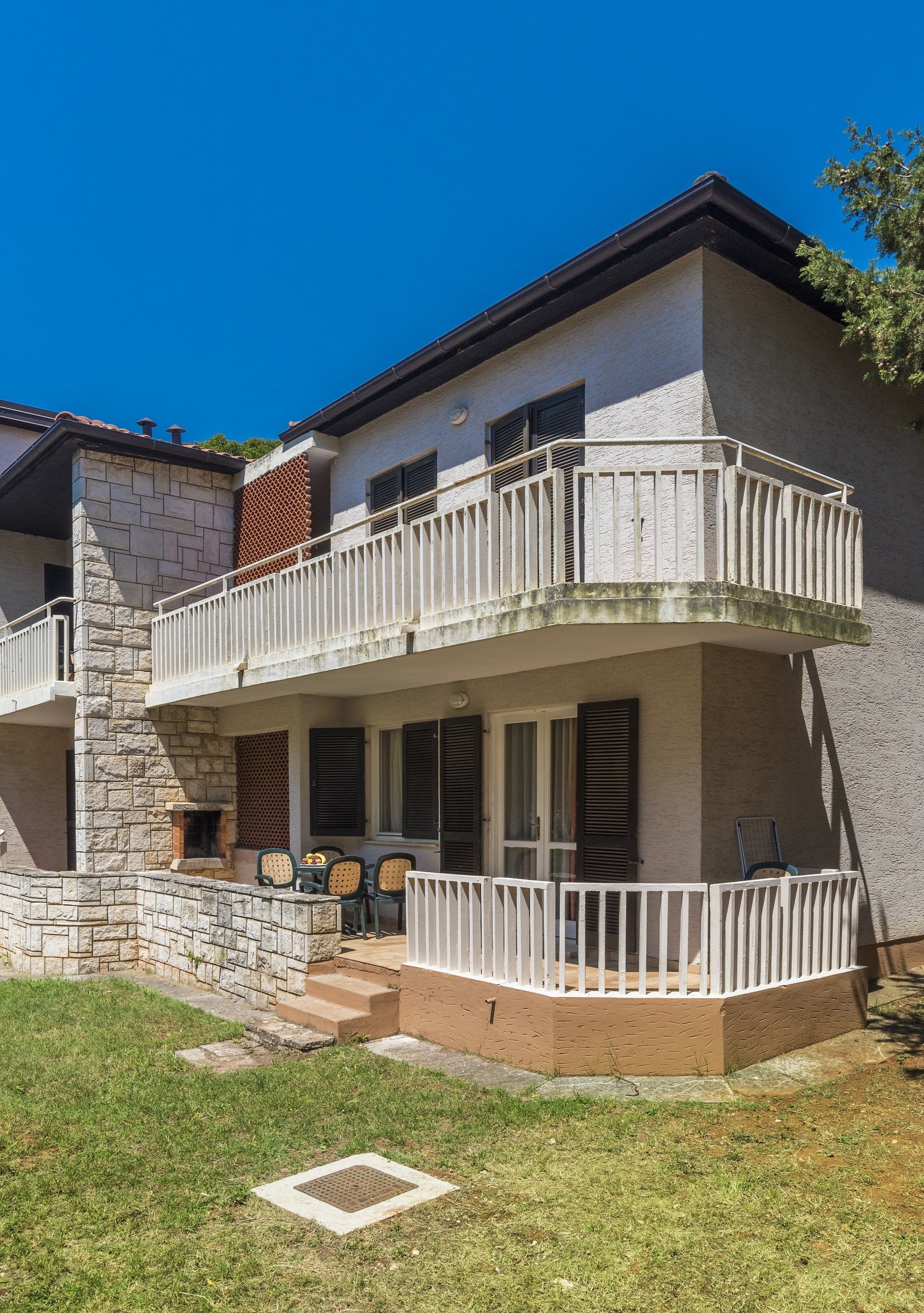 A two-story building with a stone facade, a balcony with white railings, and a small outdoor seating area with chairs and a table.