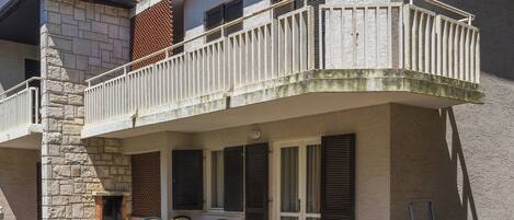 A two-story building with a stone facade, a balcony with white railings, and a small outdoor seating area with chairs and a table.
