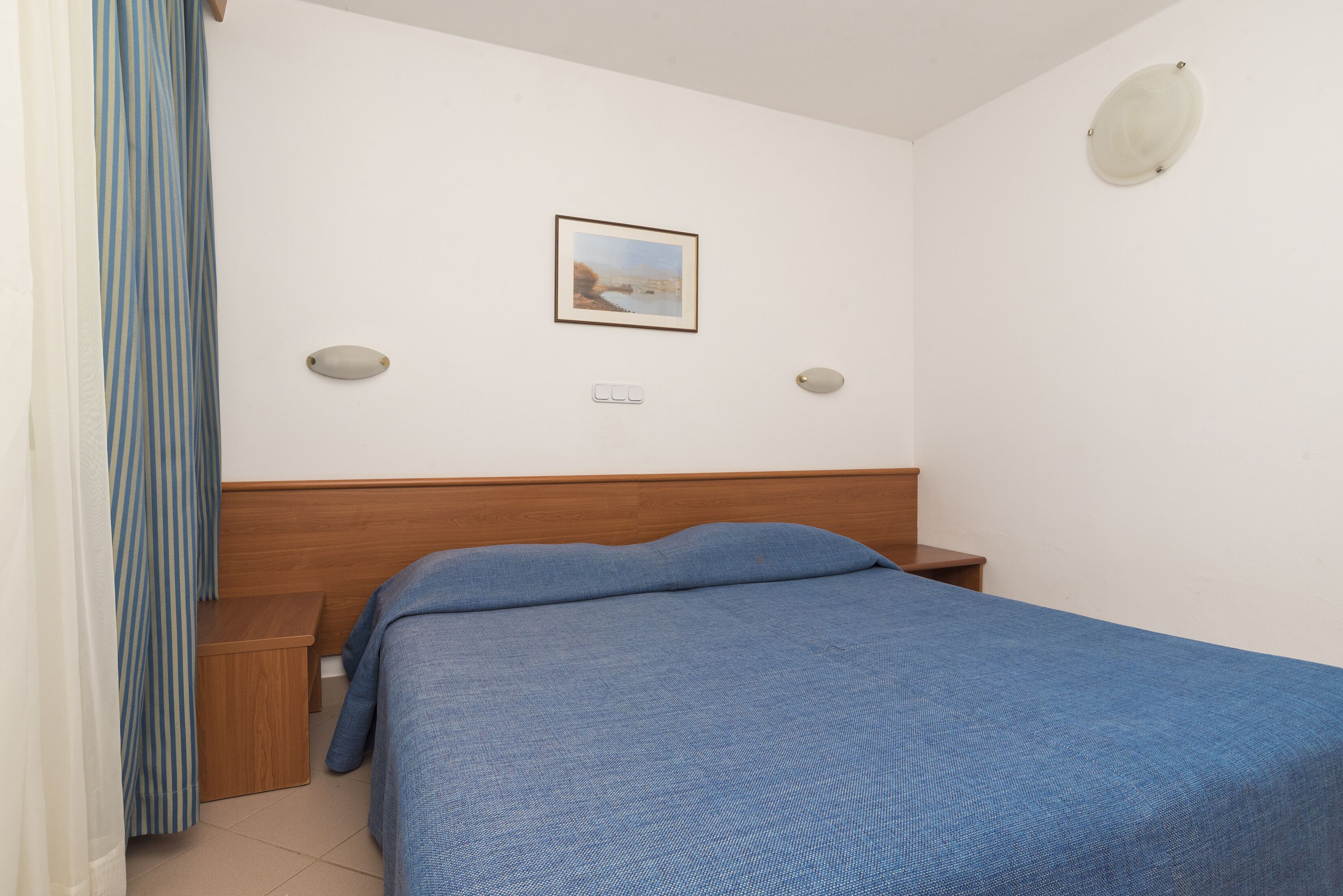 A bedroom with a wooden headboard, a single bed with a blue bedspread, a nightstand, a wall-mounted light, and a framed picture on the wall.