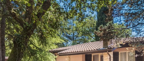 A single-story house with a tiled roof, a patio with a table and chairs, and a large tree in the foreground.