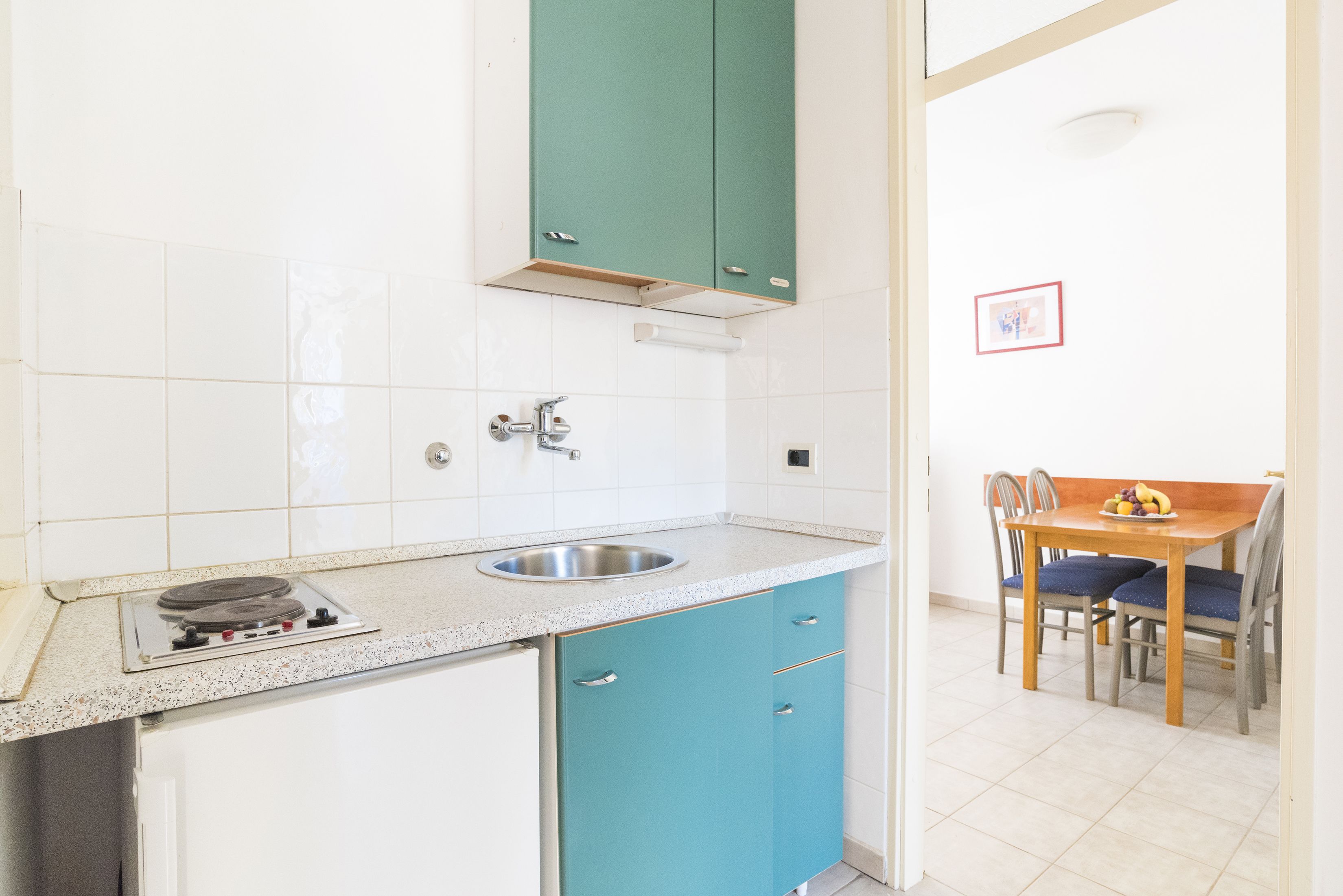 A kitchen with white tiles, a stainless steel sink, a stove, and teal cabinets.