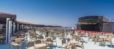 An outdoor seating area with numerous tables and chairs, a covered section with a roof, and a stage with a backdrop.