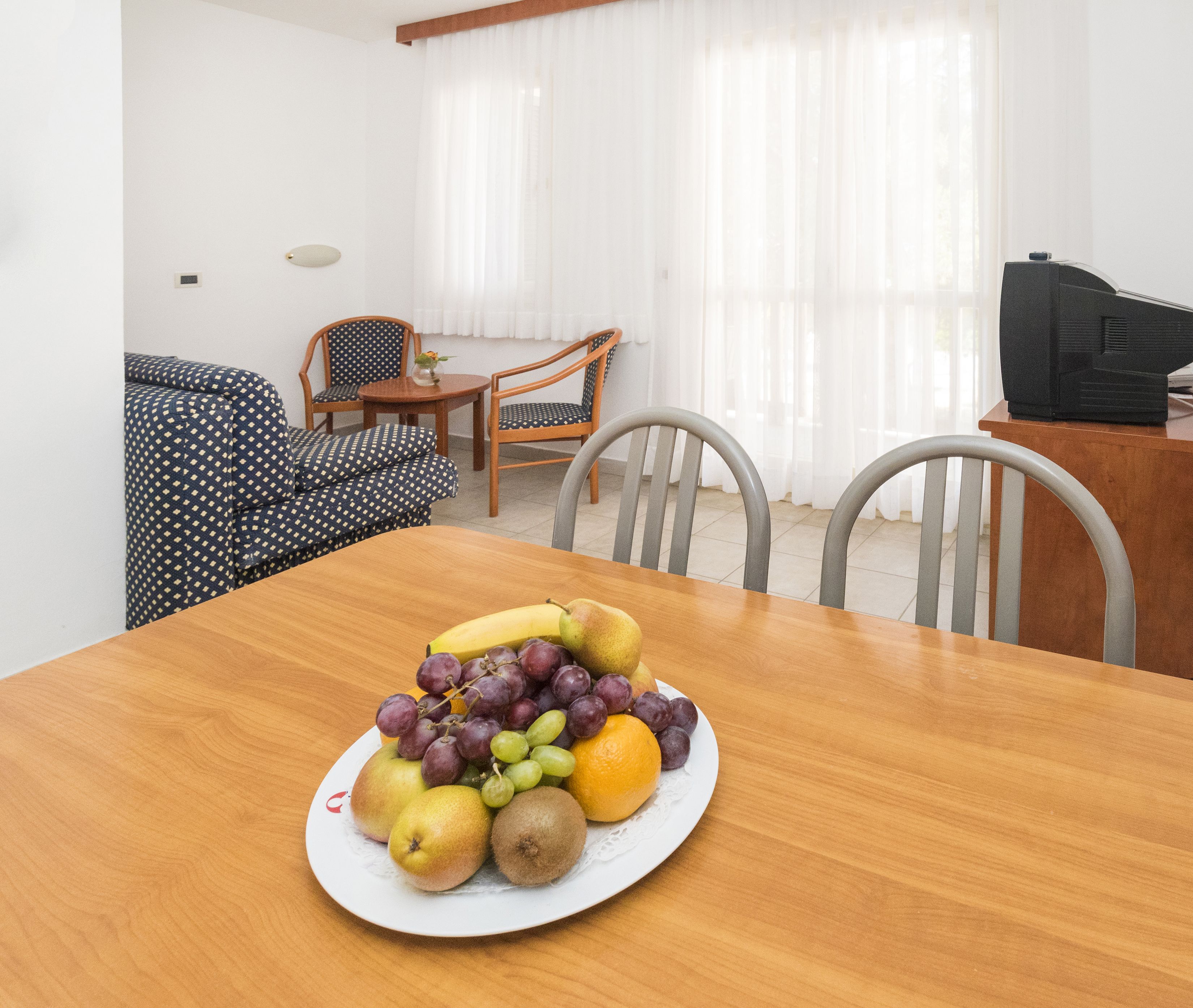 A dining table with a fruit platter, two chairs, a small table with a vase, and a television in a room with white walls and a window with sheer curtains.