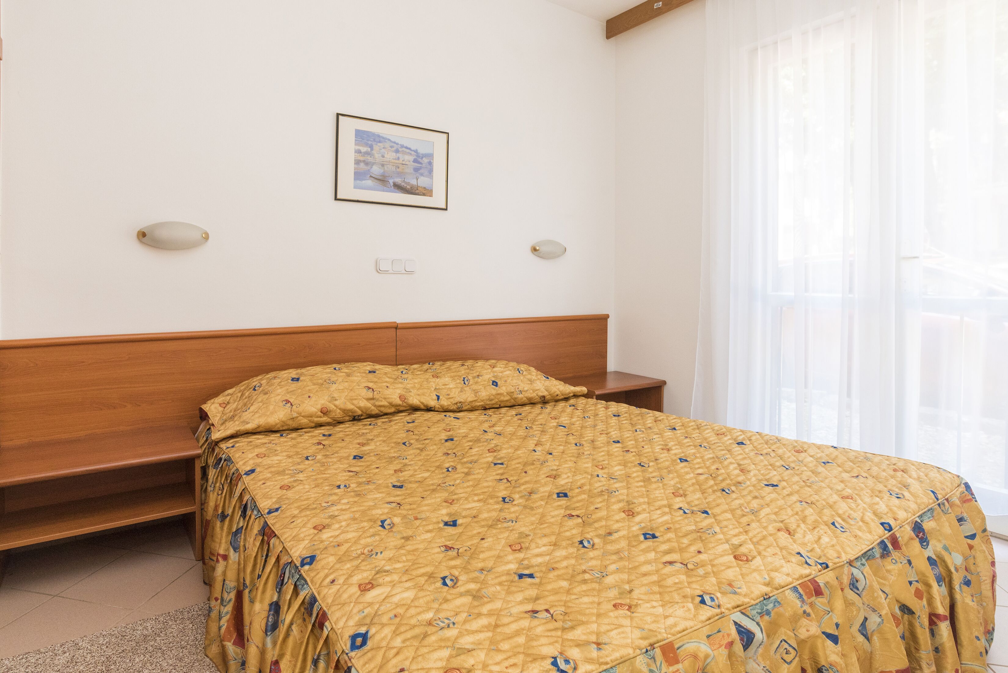 Un dormitorio con cabecera de madera, cama con edredón estampado, mesita de noche y ventana con cortinas traslúcidas.