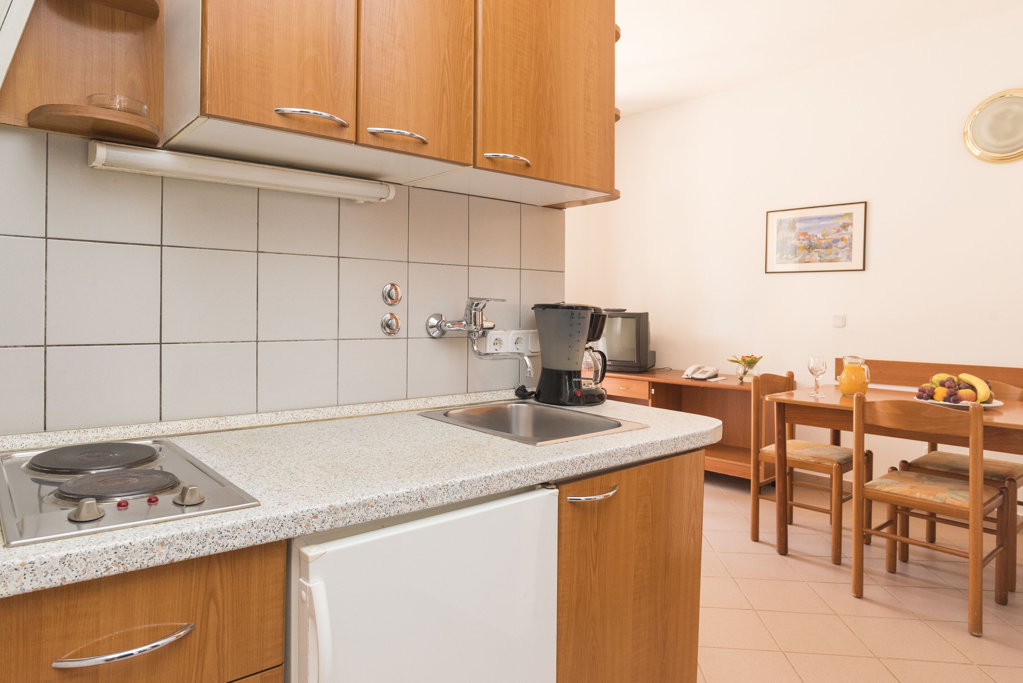 Een keuken met houten kastjes, een witte koelkast, een spoelbak en een eethoek met een tafel en stoelen.