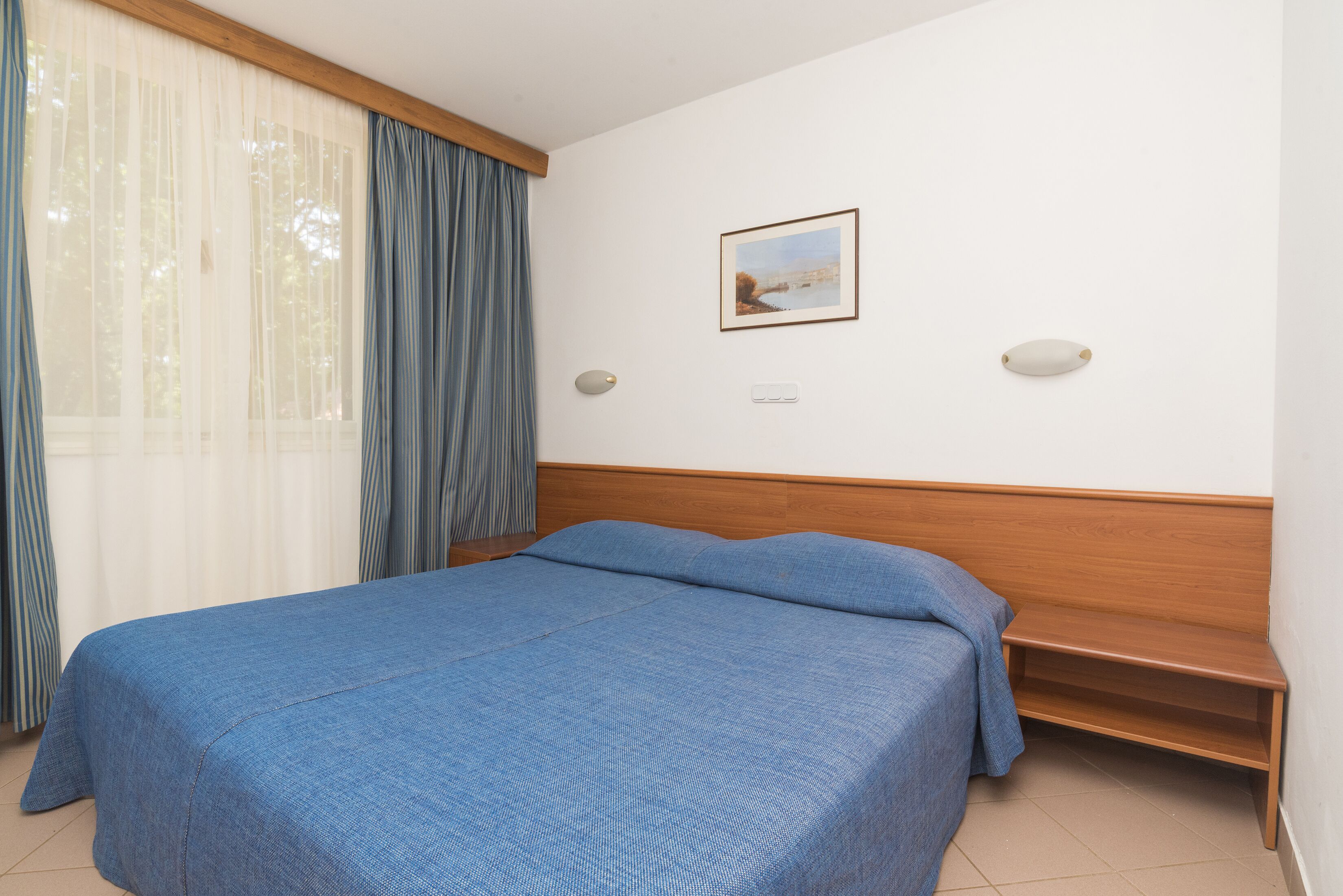 Une chambre d’hôtel avec un lit, une table de chevet, une lampe fixée au mur et un tableau encadré.