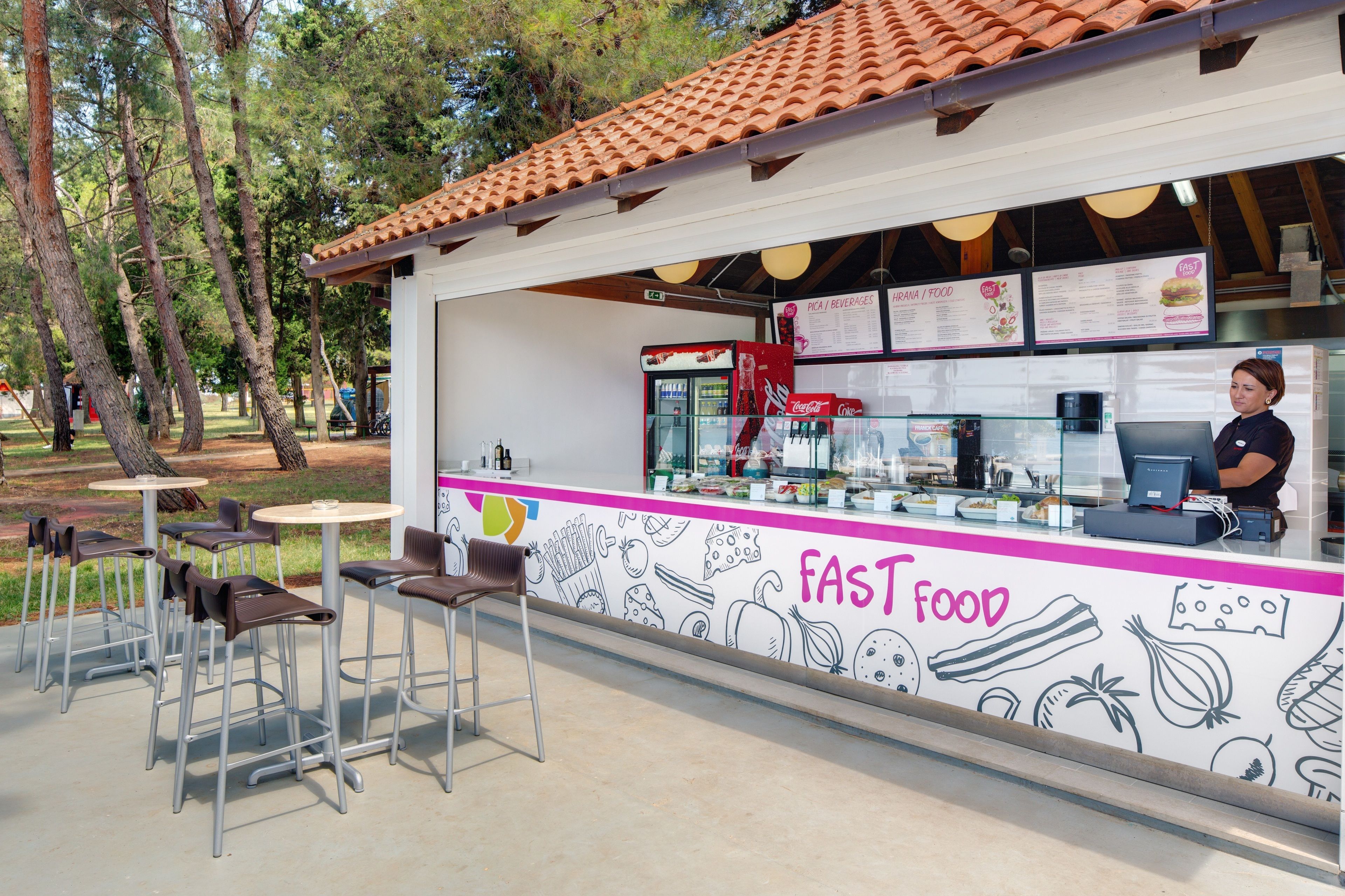 An outdoor fast food kiosk with a menu board, a Coca-Cola cooler, and a counter with a cashier.