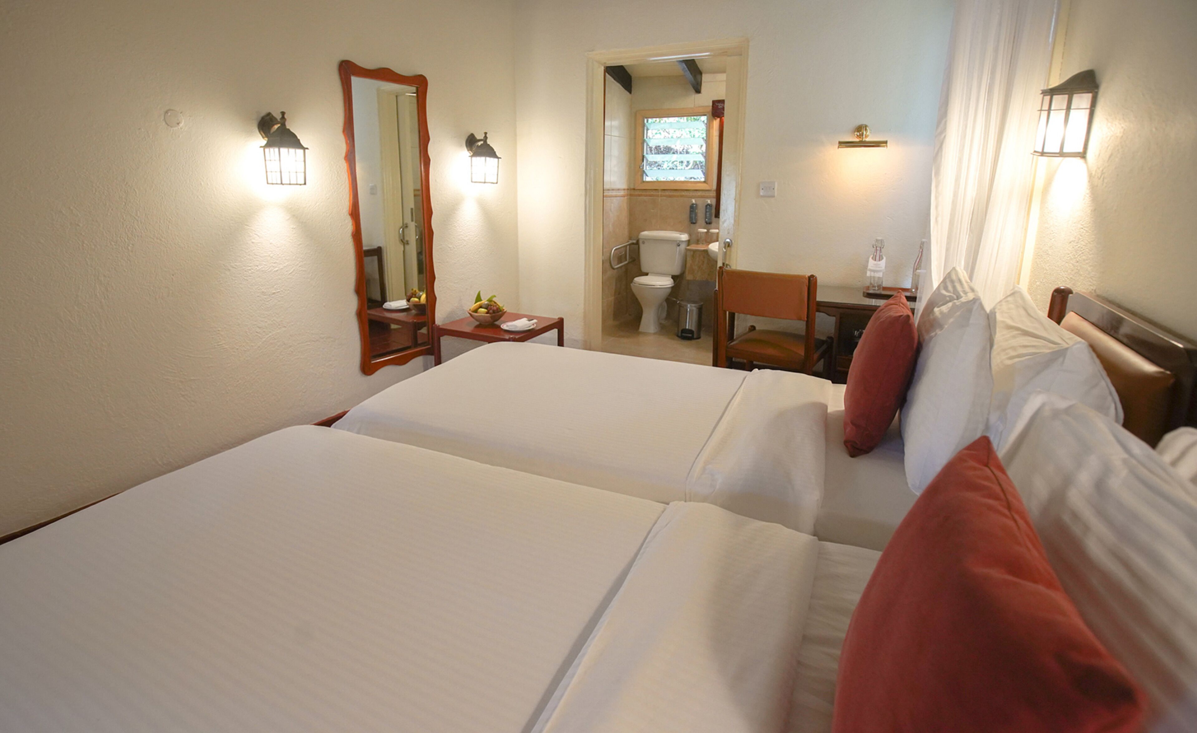 A hotel room with two beds, a small table, a chair, a mirror, and a bathroom visible through an open door.