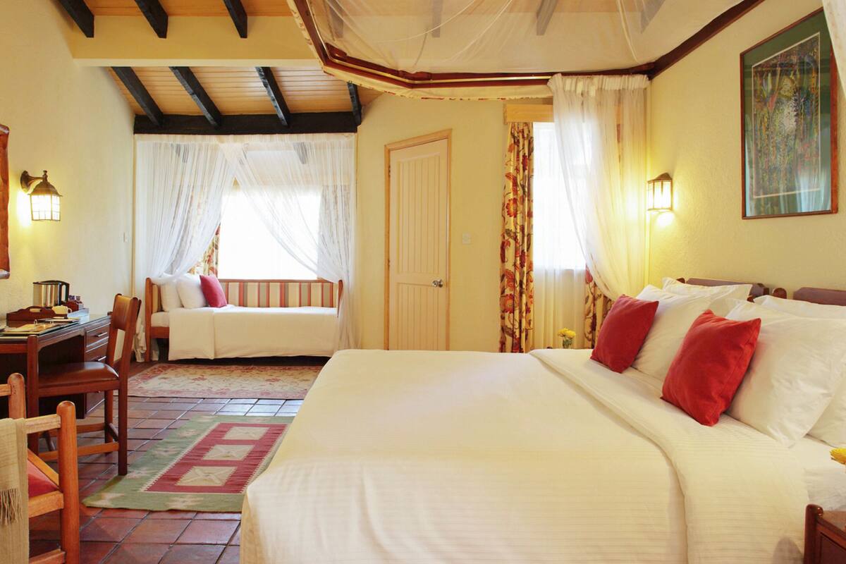 Habitación de hotel con una cama grande, un escritorio con silla, una ventana con cortinas translúcidas y un cuadro en la pared.