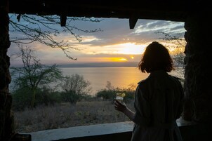 Una persona de pie junto a una ventana con vista a una puesta de sol sobre un cuerpo de agua, con árboles y un paisaje rocoso a la vista.