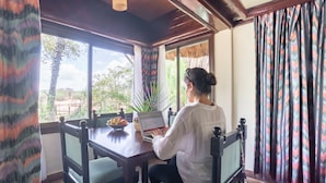 A person working at a wooden table with a laptop, near large windows with curtains, in a room with a thatched roof and wooden beams.