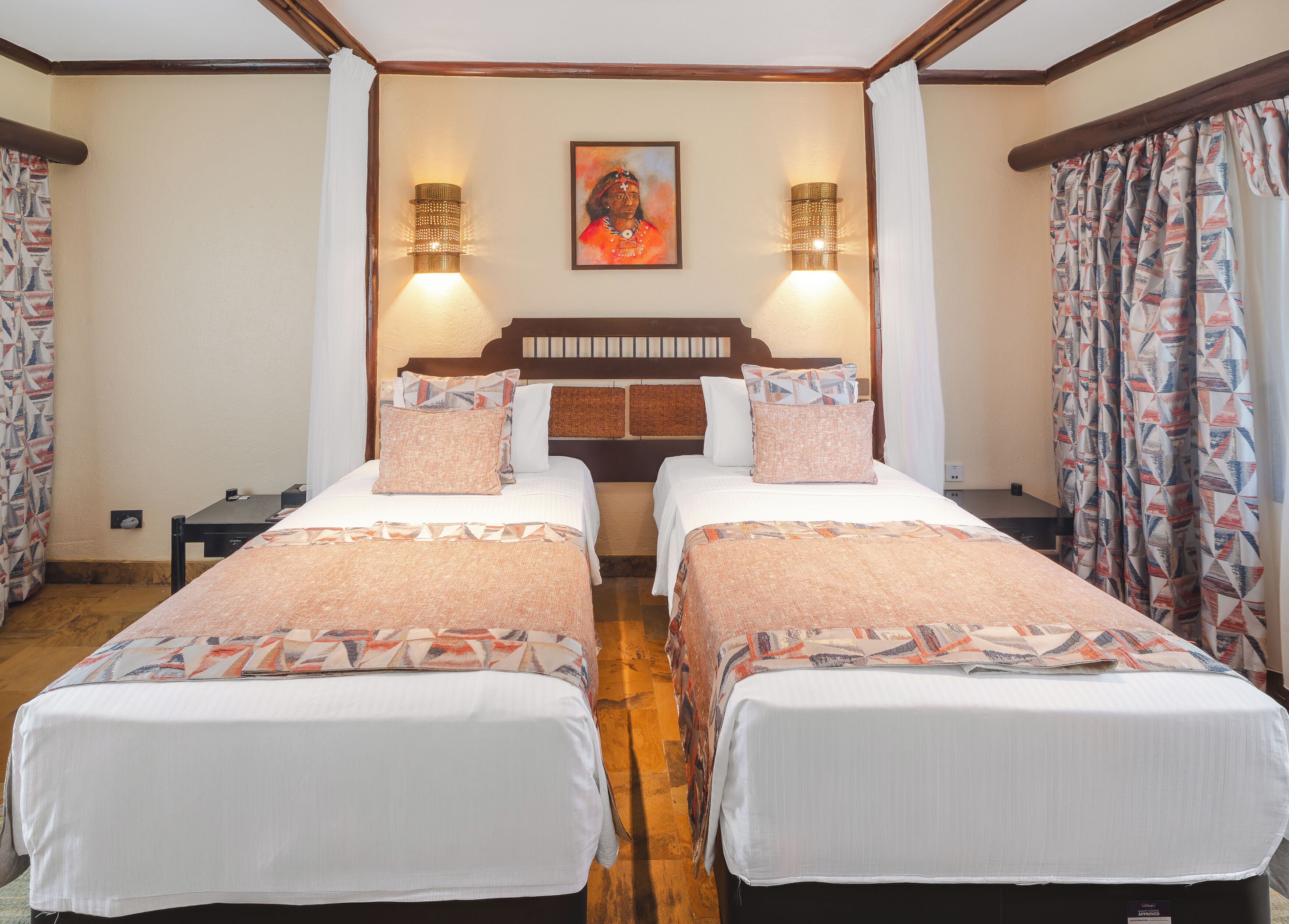 A hotel room with two beds, a wooden headboard, wall-mounted lamps, a painting, and patterned curtains.