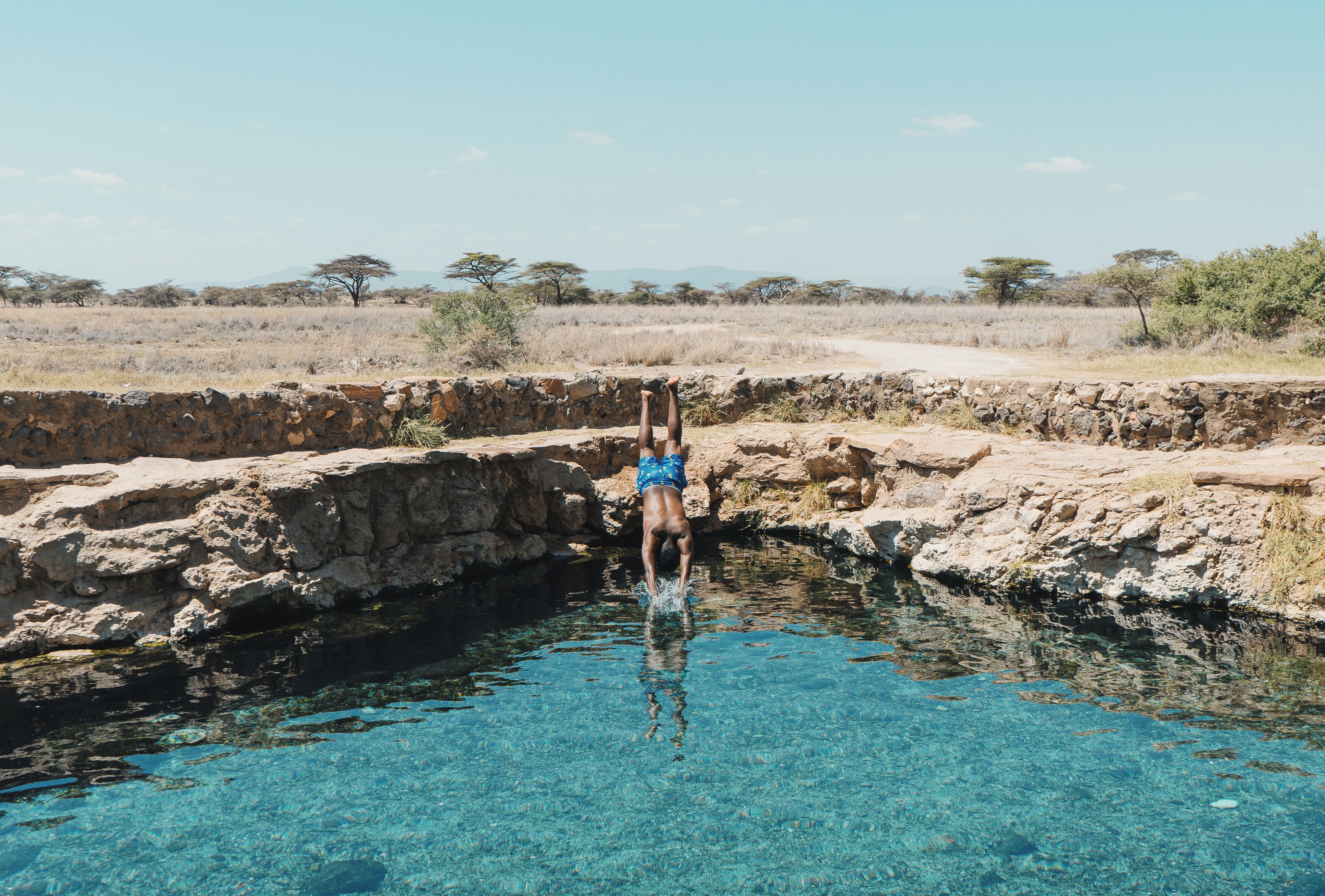 Una persona de pie en una piscina cristalina con bordes rocosos, rodeada por un paisaje árido y con escasa vegetación.