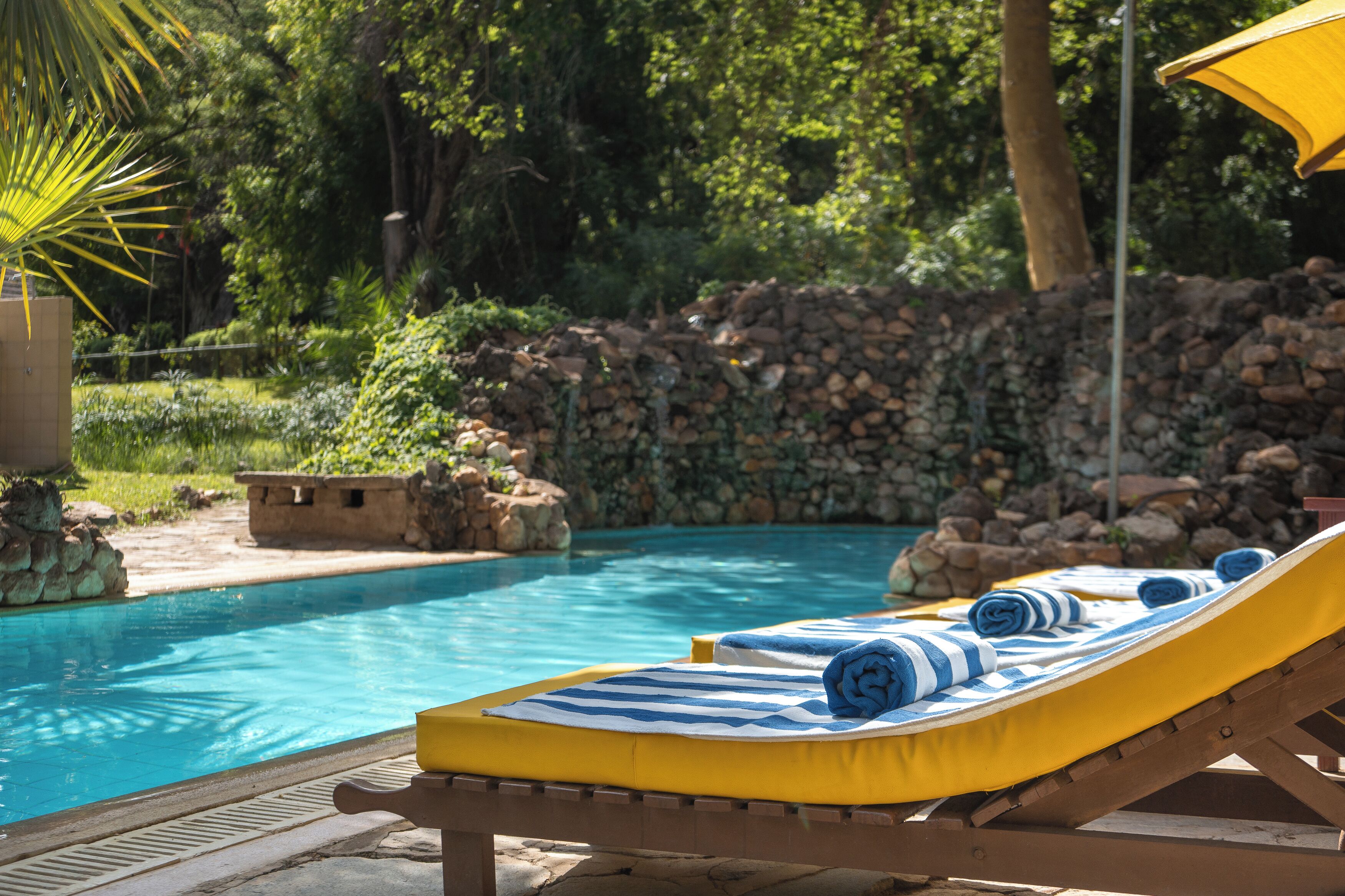Zona junto a la piscina con sillones de descanso y toallas, rodeada de vegetación y un muro de piedra.