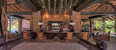 Zona de bar al aire libre con paredes de piedra, muebles de madera y techo rústico.