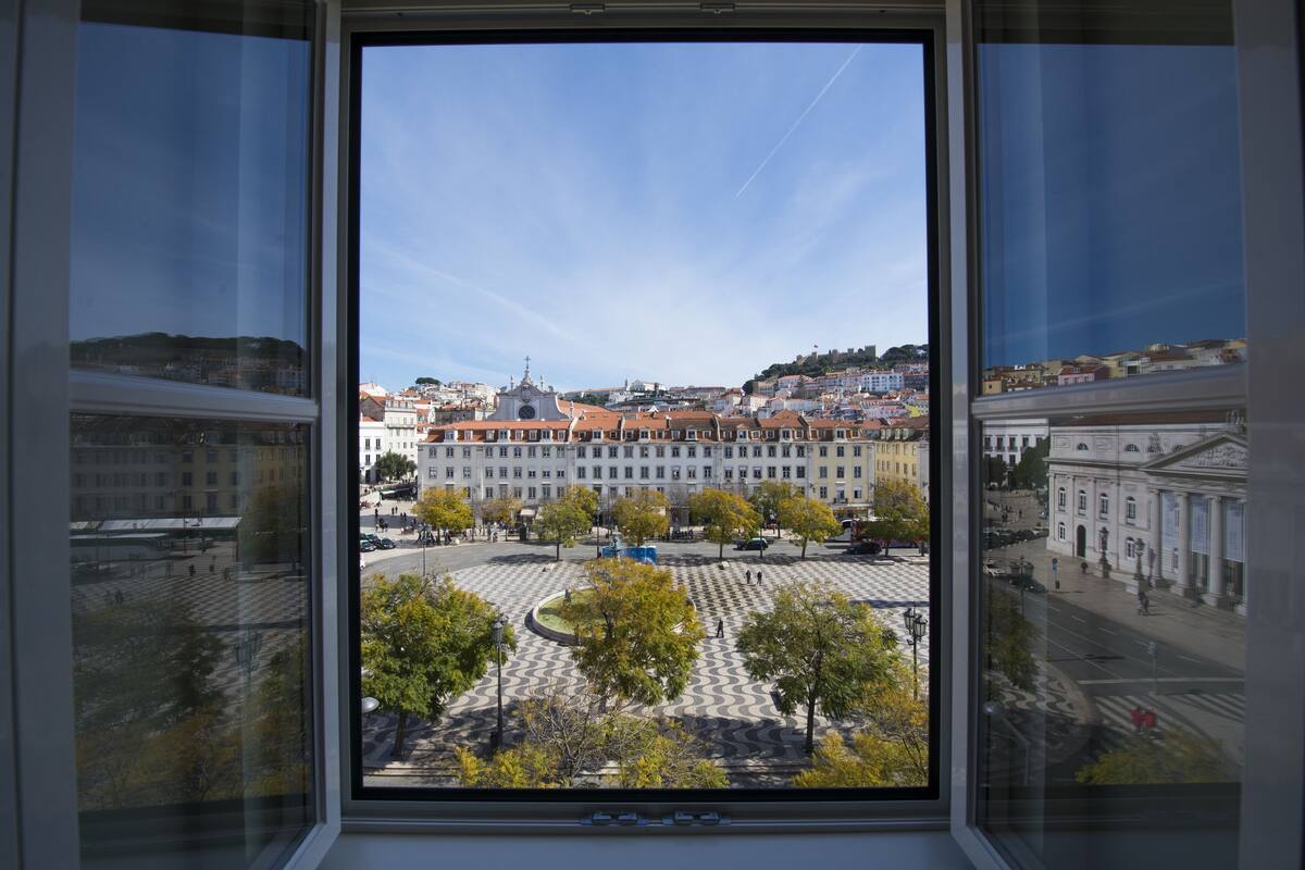 Une vue à travers une fenêtre sur une place avec un espace central ouvert, entourée de bâtiments aux toits recouverts de tuiles rouges et d’une église imposante surmontée d’un dôme.