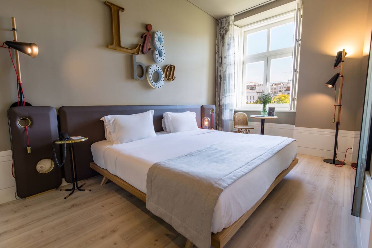 Une chambre à coucher bien rangée, avec un grand lit, des tables de chevet, une petite table sur laquelle est posé un vase, et une fenêtre avec des rideaux.