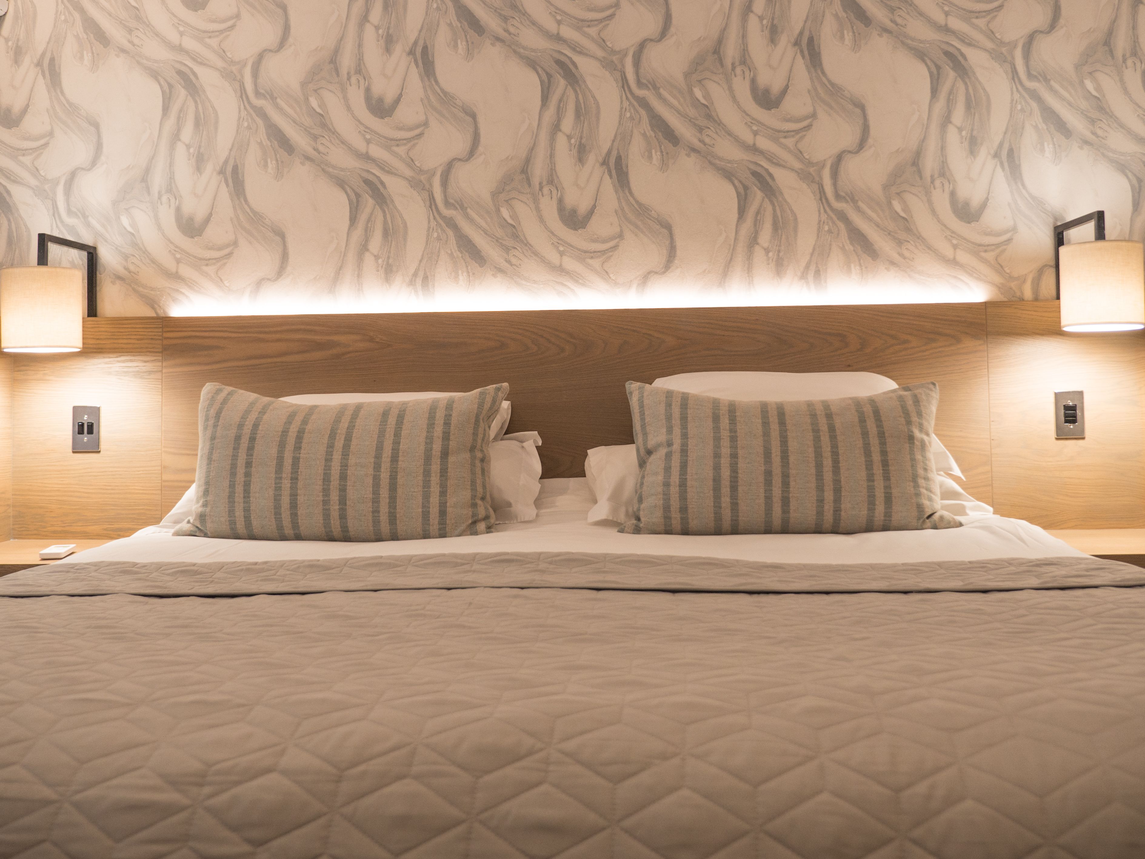 Un lit bien fait, recouvert d’une courtepointe matelassée, avec deux oreillers et une tête de lit ornée d’un motif rappelant le marbre. Deux tables de chevet sont fixées au mur, chacune étant équipée d’une lampe et d’une prise électrique.