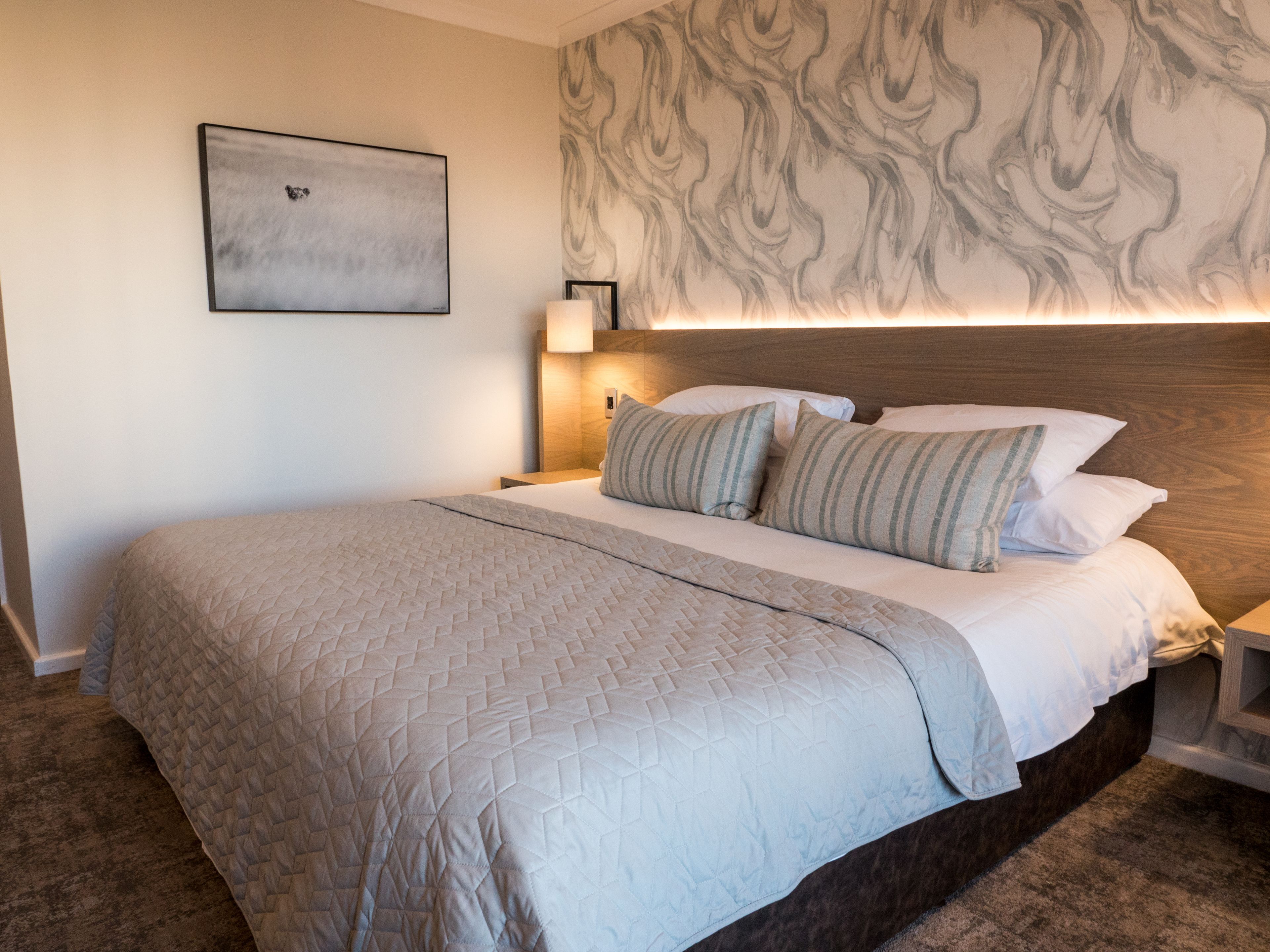A hotel room with a large bed, a headboard with a marble-like design, a bedside table with a lamp, and a framed picture on the wall.