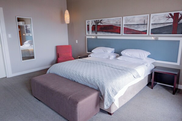 A bedroom with a bed, a bench, a chair, a bedside table, and wall art. - The Kelway Hotel (Gqeberha (Port Elizabeth))