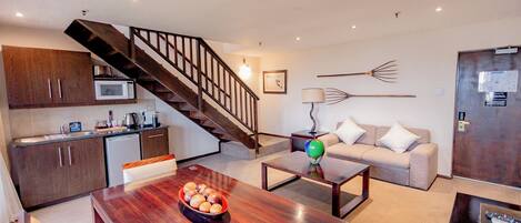 A modern kitchen with a microwave, a dining table with a bowl of fruit, a staircase, a sofa, and a lamp.