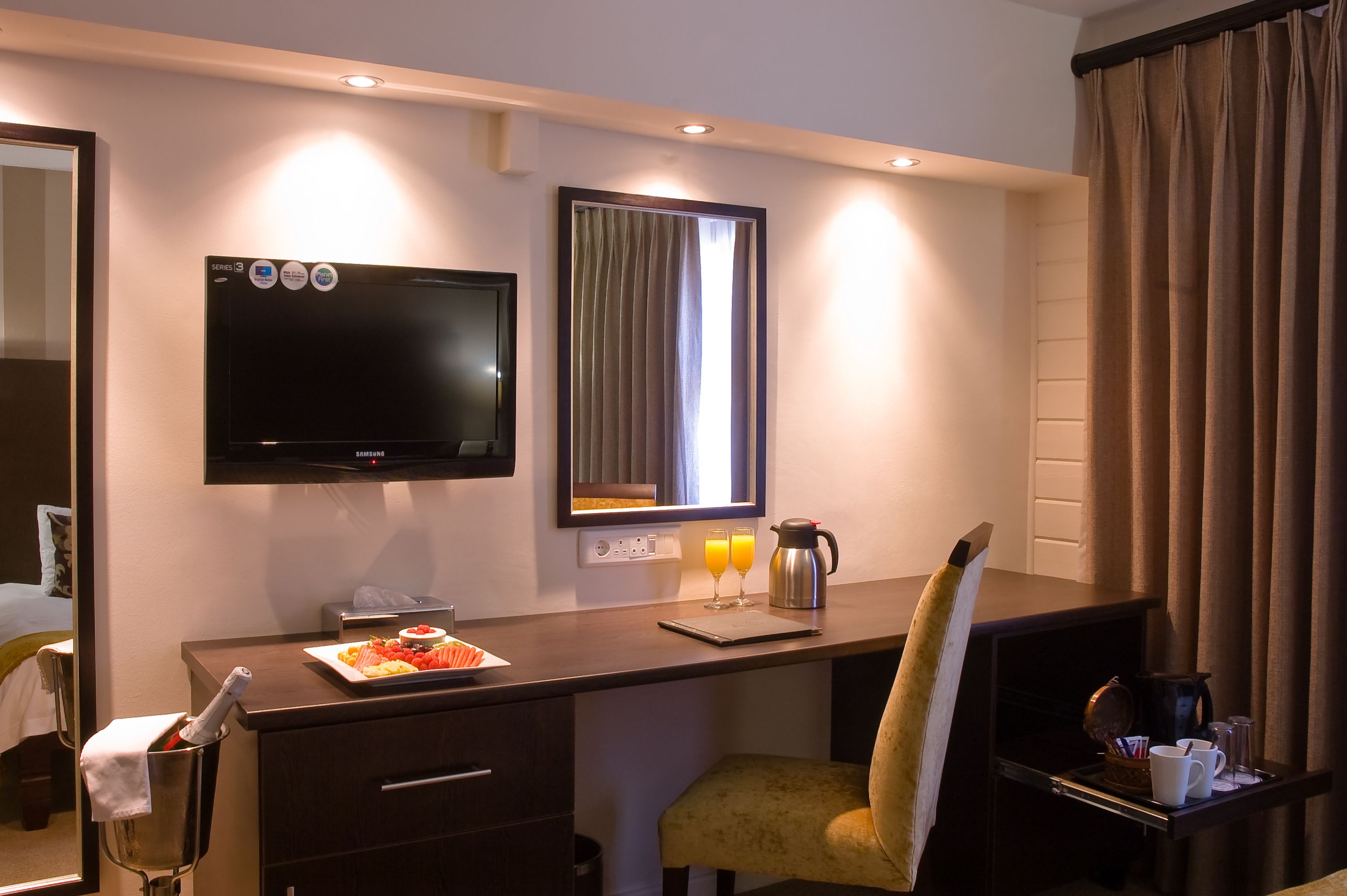 A hotel room with a bed, a desk with a flat-screen TV, a chair, a small table with a pitcher of orange juice, and a window with curtains.