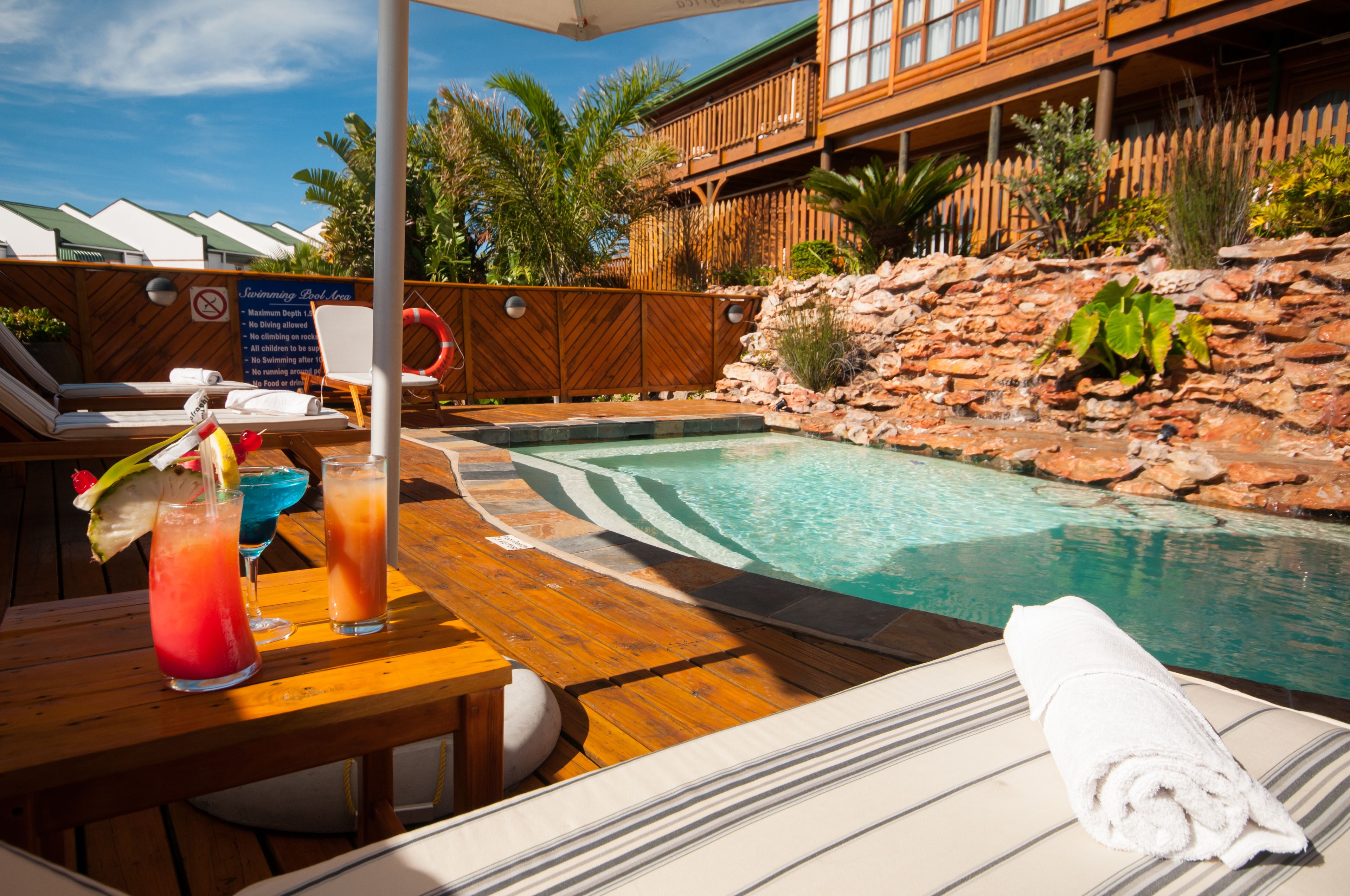 A poolside area with a wooden deck, lounge chairs, a table with drinks, and a view of a building with a wooden deck and a rock wall.