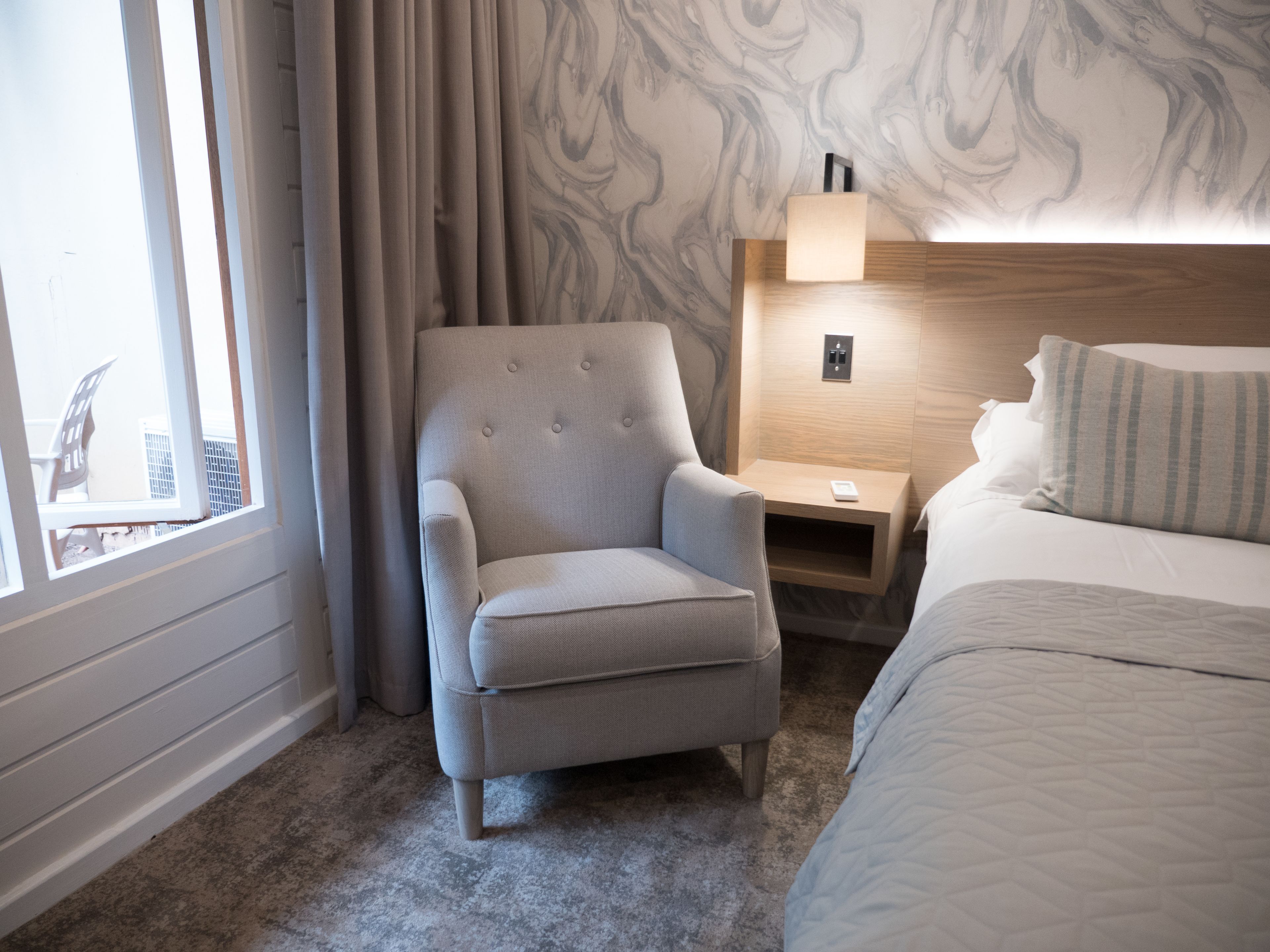 A hotel room with a large window, a grey armchair, a bedside table with a lamp, and a bed with a striped pillow.