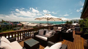 Eine Holzterrasse mit Gartenmöbeln, darunter Sofas und ein Tisch, mit Blick auf einen Strand und das Meer.
