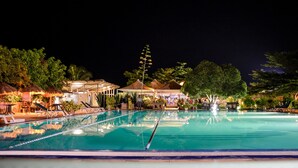 Ein Pool bei Nacht mit klarem Wasser, umgeben von Liegestühlen und Sonnenschirmen, mit Bäumen im Hintergrund.
