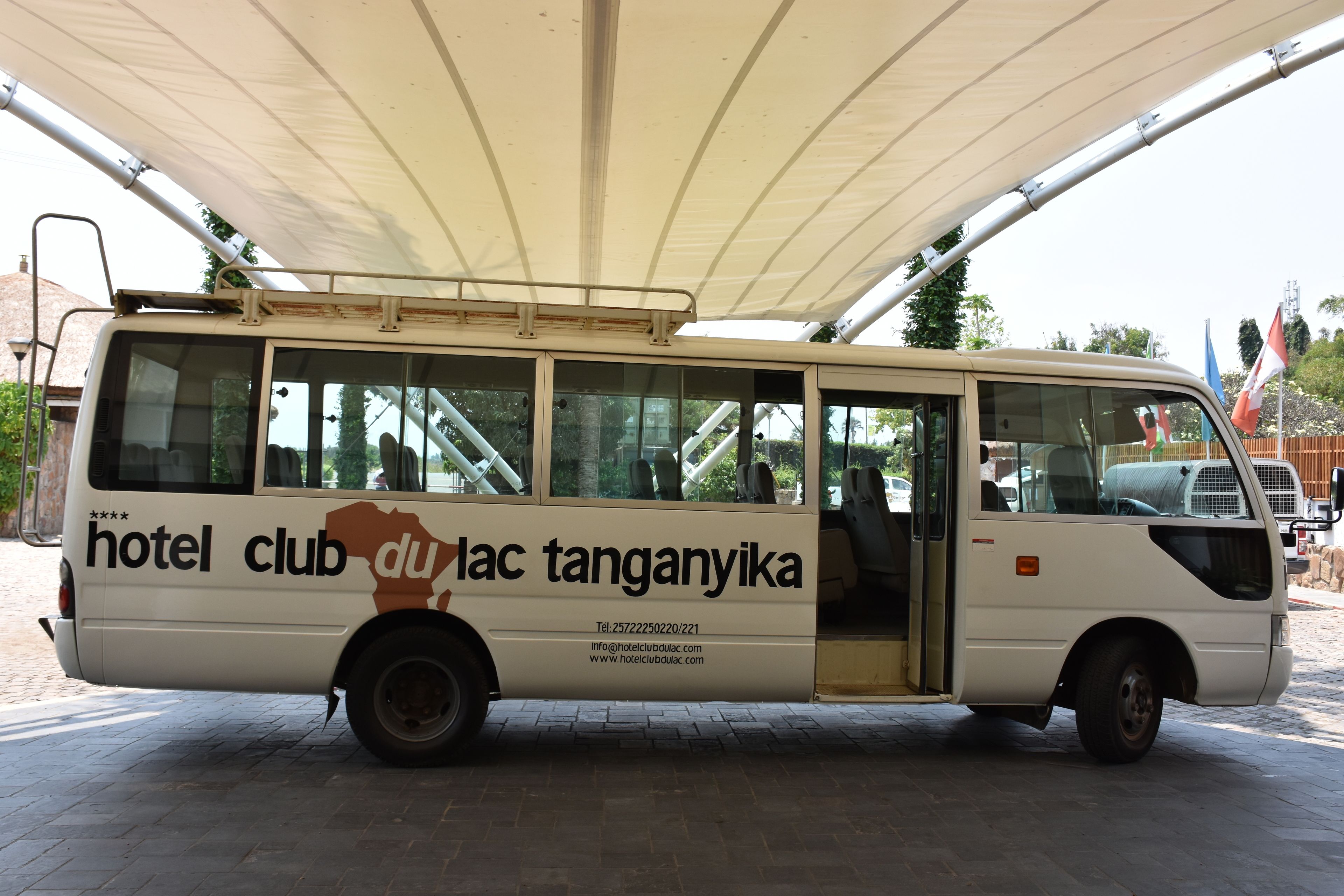 A white bus with "hotel club" and "dulac tanganatika" written on the side, parked under a canopy.